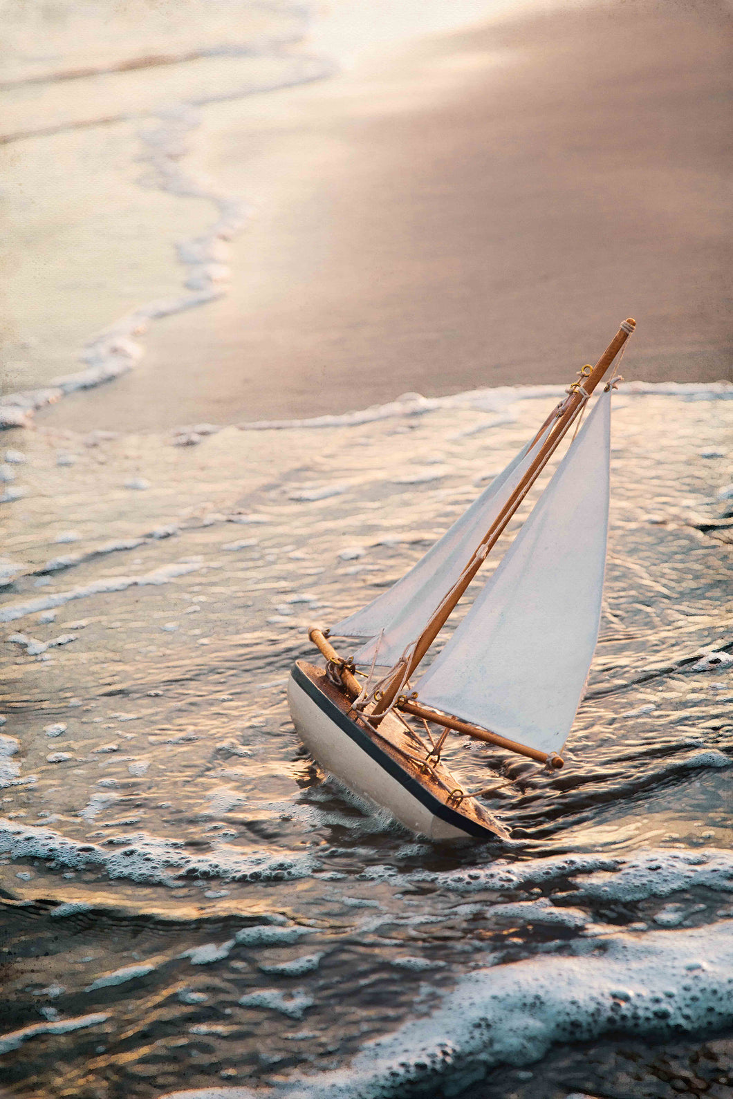 fine art mixed media image of vintage toy sailboat left on the beach with soft waves